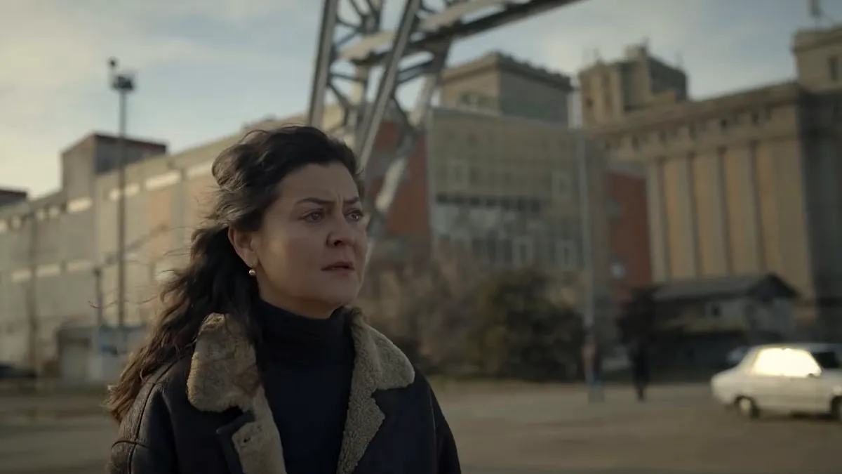 A woman in a shearling coat stands in an industrial yard before grain silos and a steel gantry, a white car passing behind her.