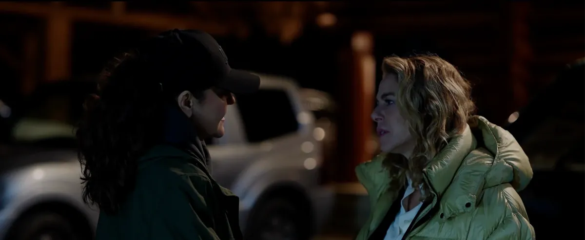 At night beside a white pickup truck, a woman in a black cap and dark green parka faces a blonde woman in an olive puffer jacket.