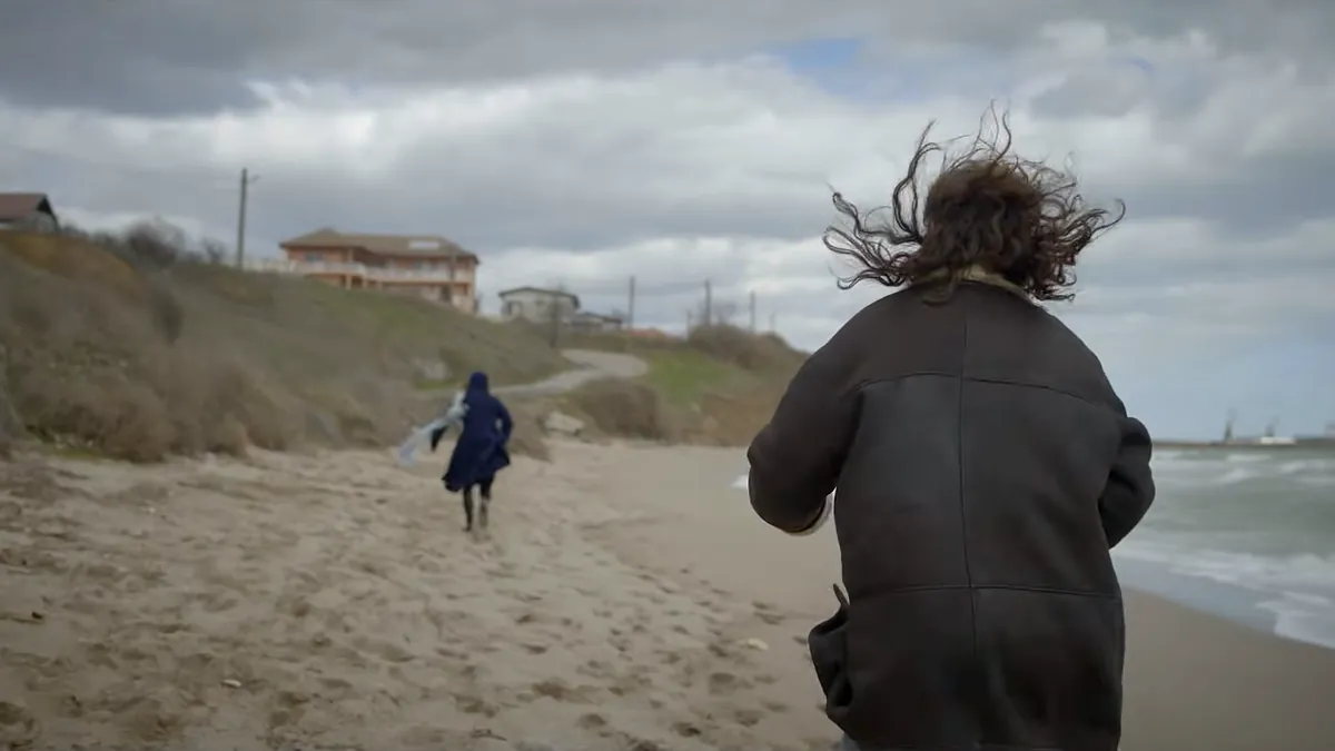 A woman in a sheepskin coat runs along a windswept beach after a distant figure in dark blue, under a low gray sky.