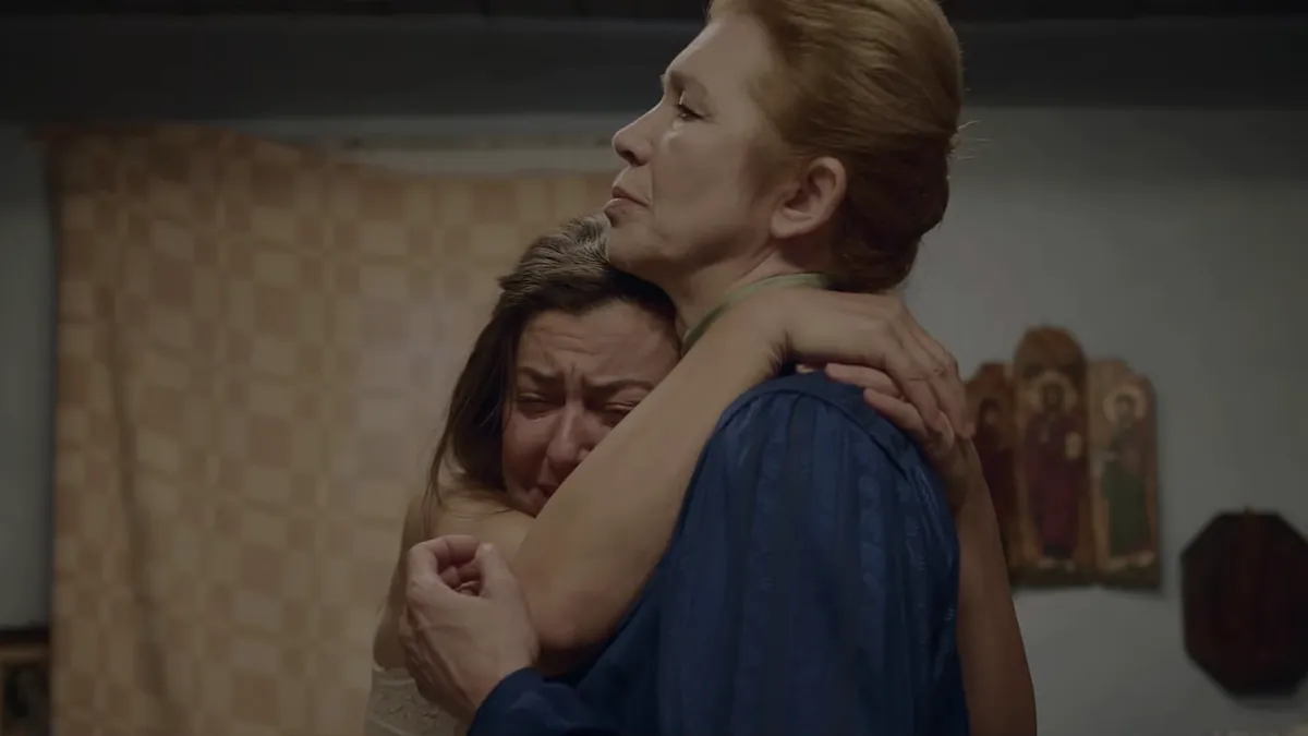 An older woman holds a weeping younger woman in her arms, icons on the wall behind them.