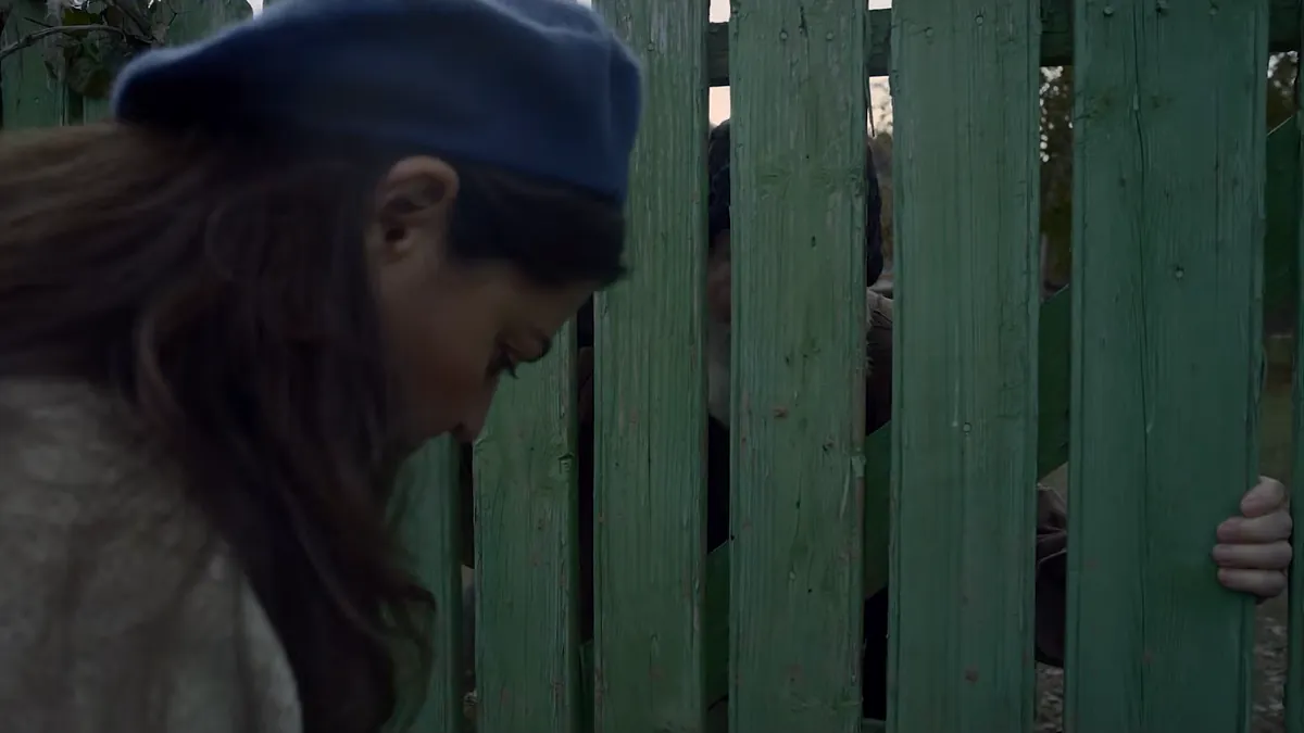 A woman in a blue beret bows her head at a green wooden fence; someone stands on the other side of the boards.