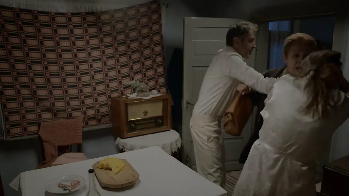 In a room with a woven wall carpet and an old radio, a man in a white shirt struggles with two women over a leather bag.