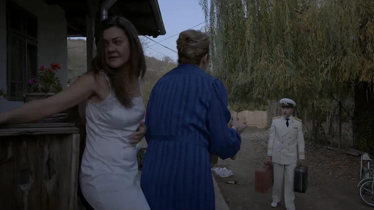 On a village porch an older woman in a blue housecoat holds back a young woman in a white slip, while a man in a white naval uniform walks up the lane with two suitcases.