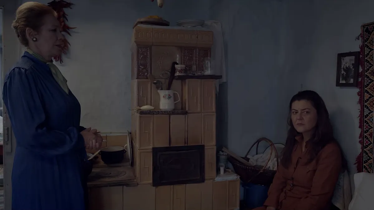 In a village room with a tiled stove, an older woman in a blue dress stands while a younger woman sits on a bench and looks at her.