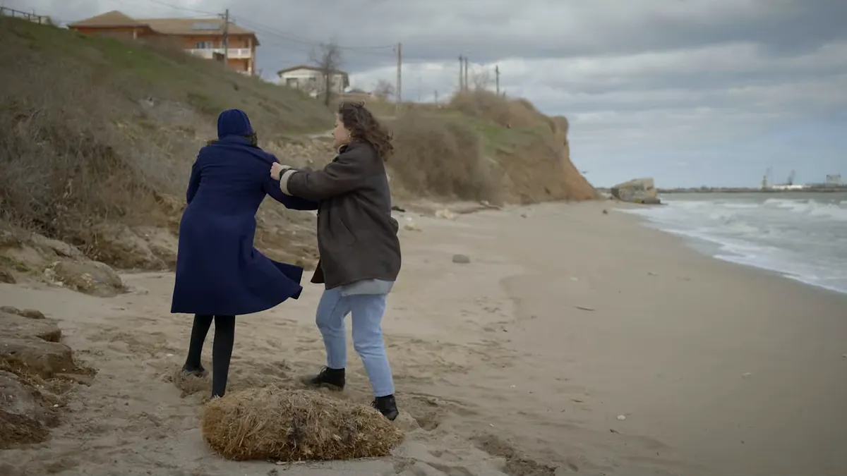 Two women grapple on a beach below a crumbling cliff, the sea gray behind them.
