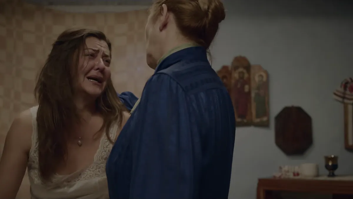 A young woman in a lace slip sobs as an older woman in a blue housecoat faces her, icons on the wall behind them.