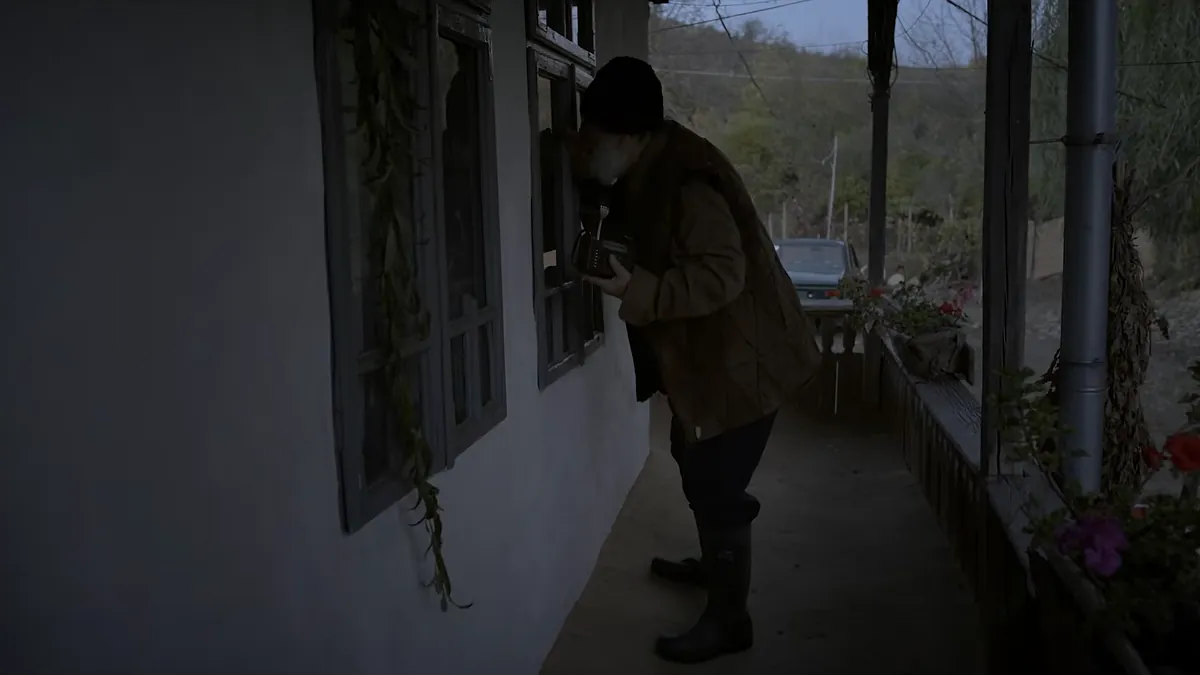 On a village porch a man in a cap and a brown jacket peers in through a window, wooded hills behind the house.