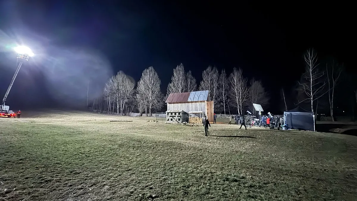Night shoot: a light on a crane floods a frosty field with a wooden hut, bare birch trees, and the crew’s tents.
